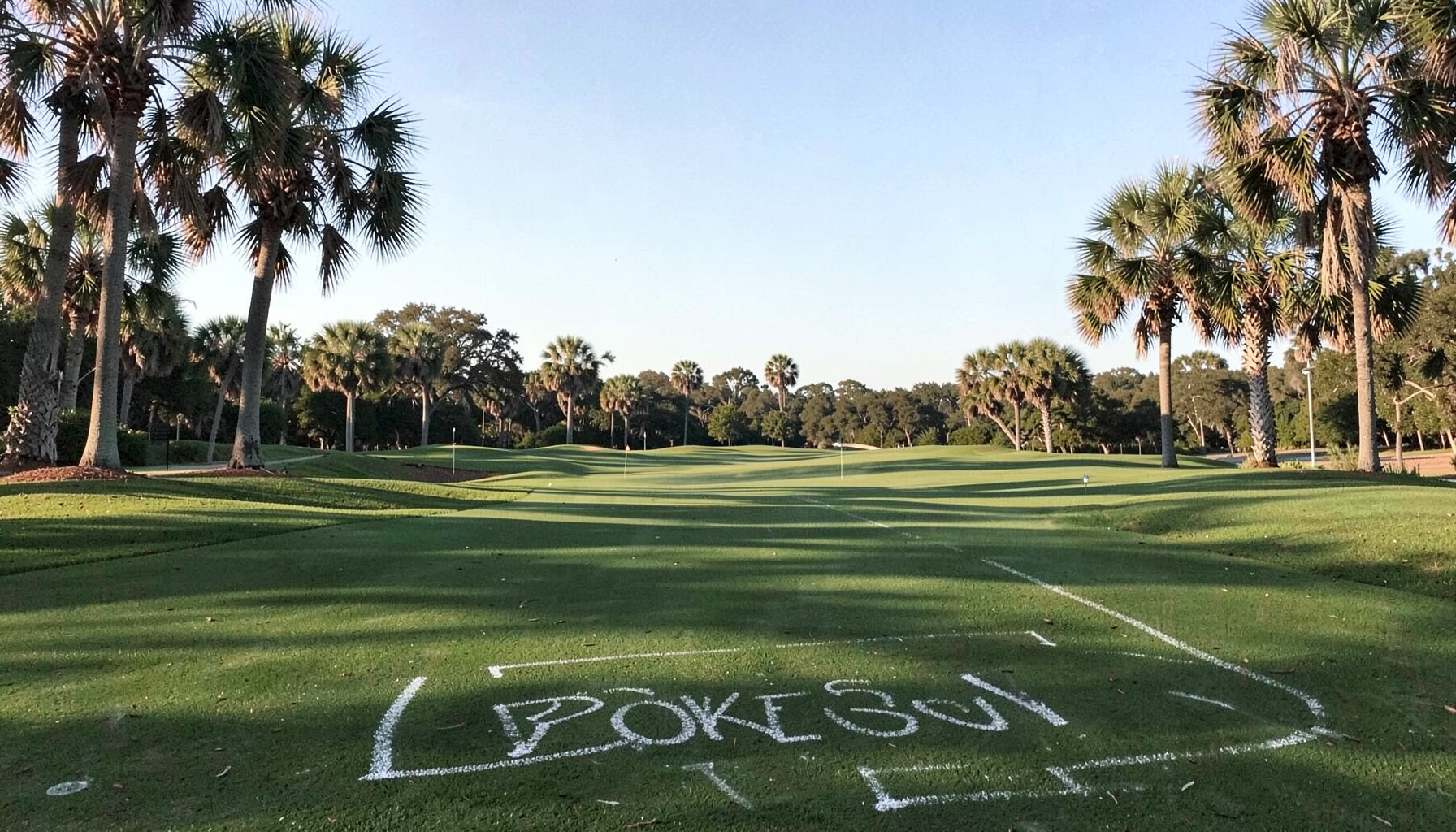 Sunlit golf fairway on The Old Course at Broken Sound, framed by mature palms and a pale blue Florida sky, with soft morning shadows stretching across manicured green turf.